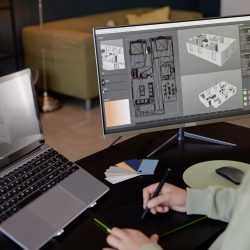 Individual engrossed in creating detailed architectural design on computer using modern digital tools, with another laptop on side for reference and color samples laying on desk